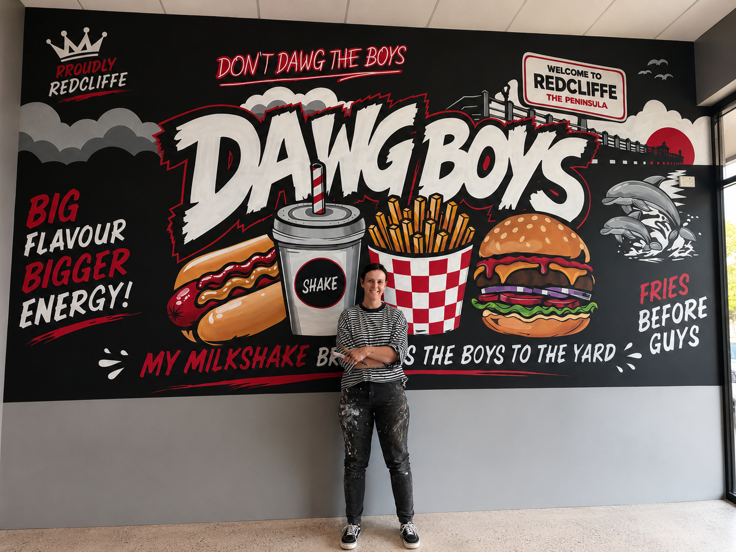 Finished DawgBoys mural in Redcliffe, bold black, white and red hand-painted lettering with a burger, hot dog, milkshake and fries, with the artist standing in front